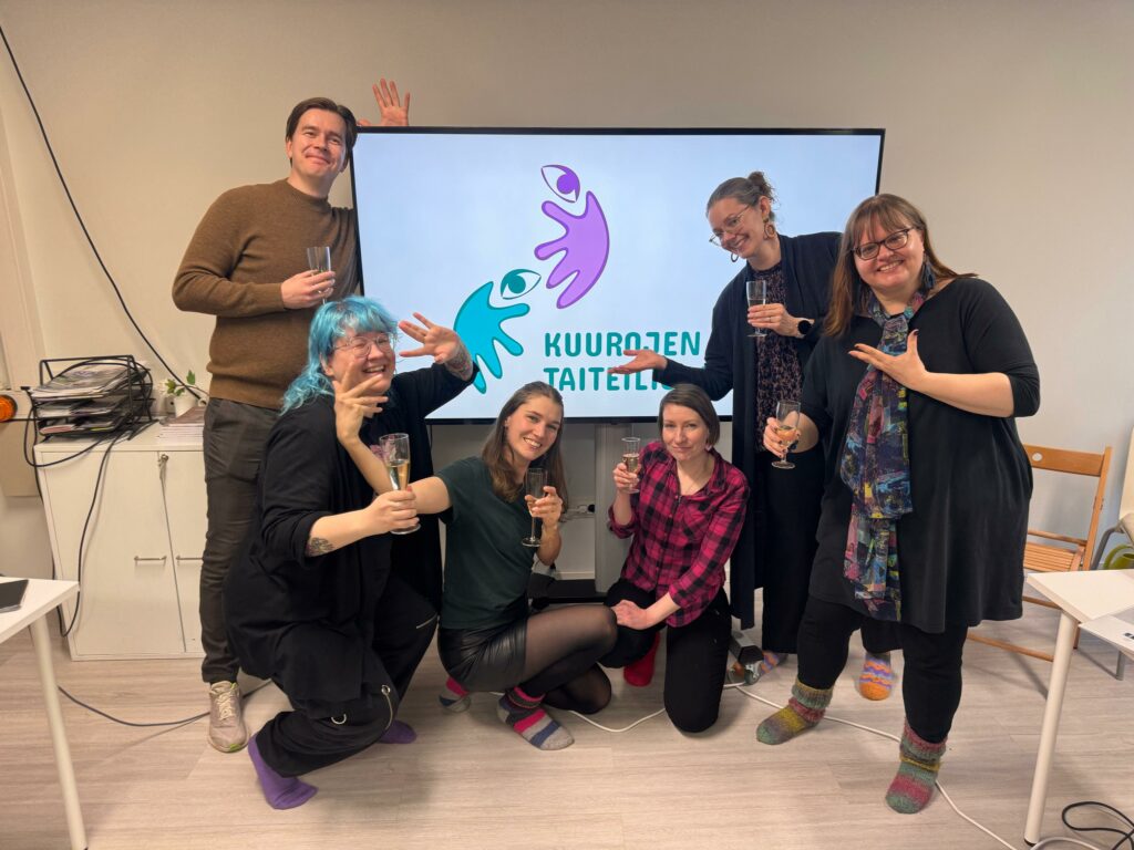 Kuurojen Taiteilijakillan hallitus poseeraa. Kuusi iloista henkilöä skumppalasit kädessään. Taustalla televisioruutu, jossa on Taiteilijakillan uusi logo ja teksti Kuurojen taiteilijakilta.